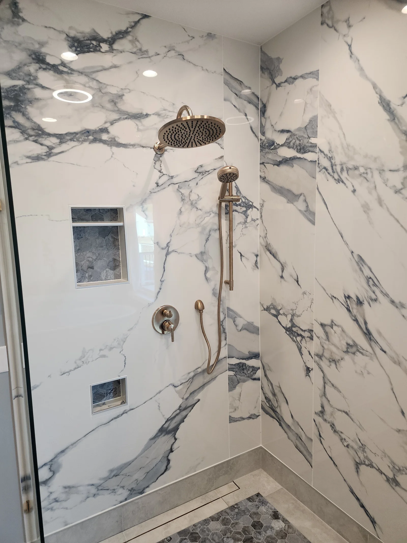 Walk-in shower with large marble-look wall panels, brushed brass rain showerhead and handheld spray, and gray hex tile niches