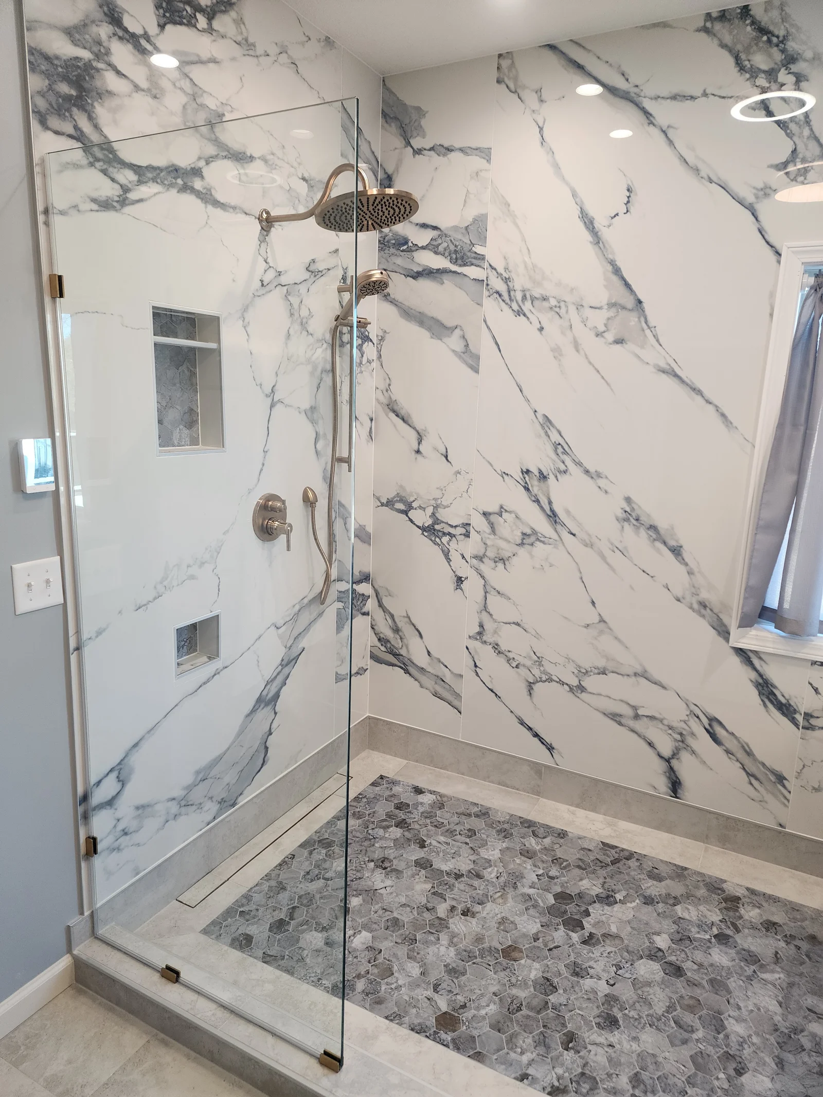 Walk-in shower with marble-look large-format wall tile, gray hexagon mosaic floor, frameless glass door, and brushed brass fixtures