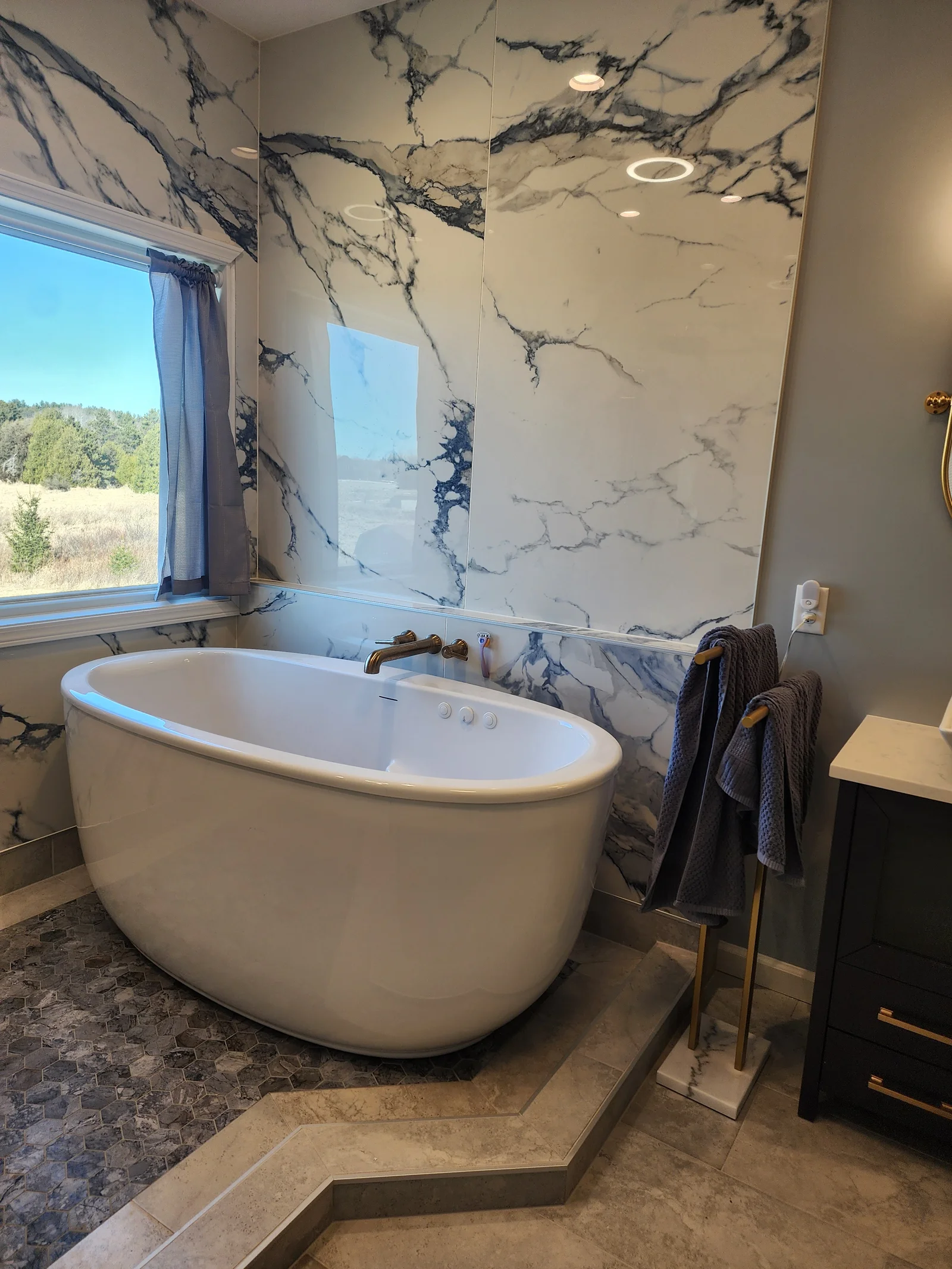 Bathroom with freestanding white tub, marble-look wall panels, hexagon mosaic floor, and brushed gold fixtures