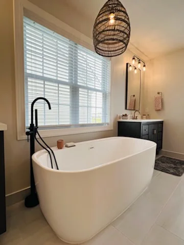 Freestanding white soaking tub with matte black floor filler beside a window, large tile floor, and dark gray vanity