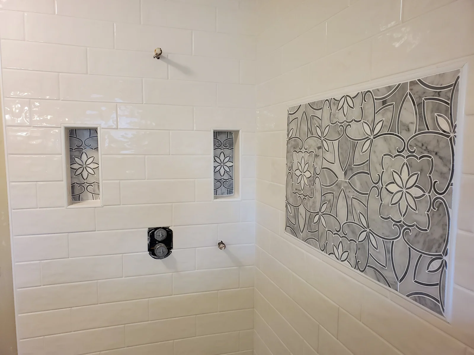 Shower wall with white glossy subway tile, recessed niches with gray marble floral mosaic inserts, and a large marble mosaic accent panel