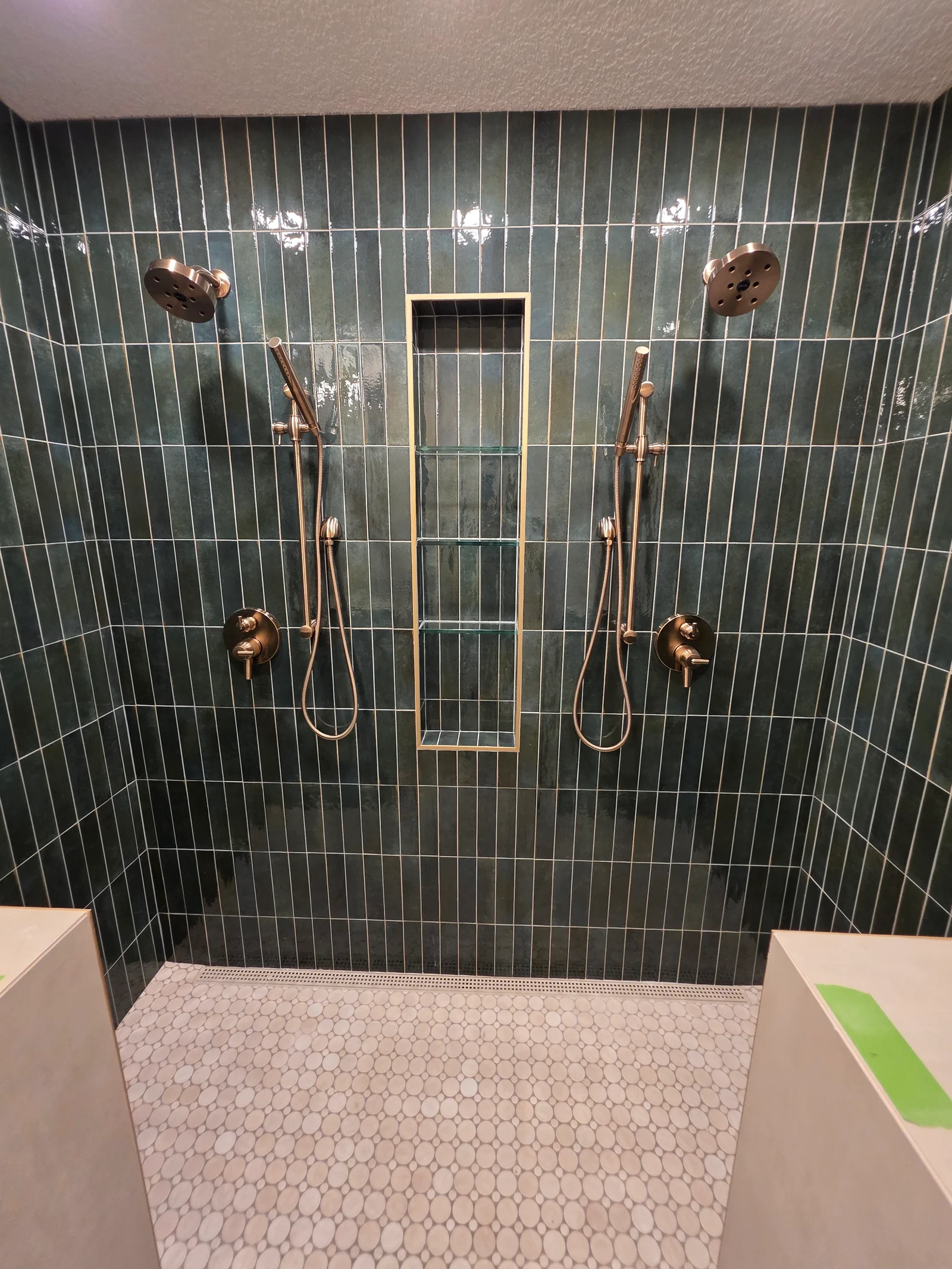 Walk-in shower with dark green glossy vertical tile, brushed gold dual shower heads, handheld sprayers, recessed niche with glass shelves, and white pebble mosaic floor