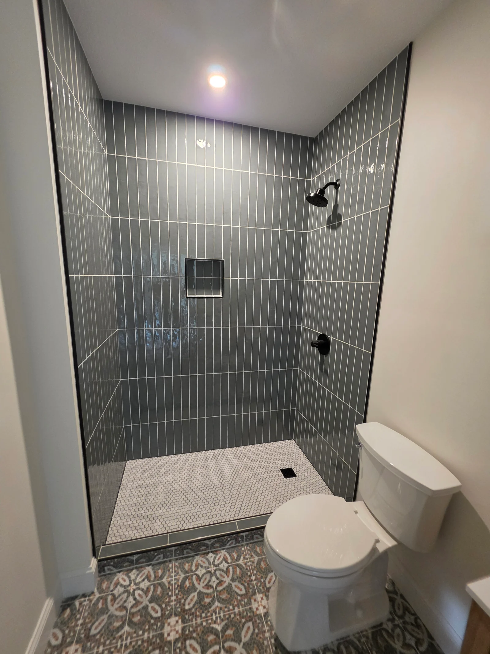 Walk-in shower with dark gray vertical glossy subway tile, white hex mosaic shower floor, recessed niche, and matte black fixtures